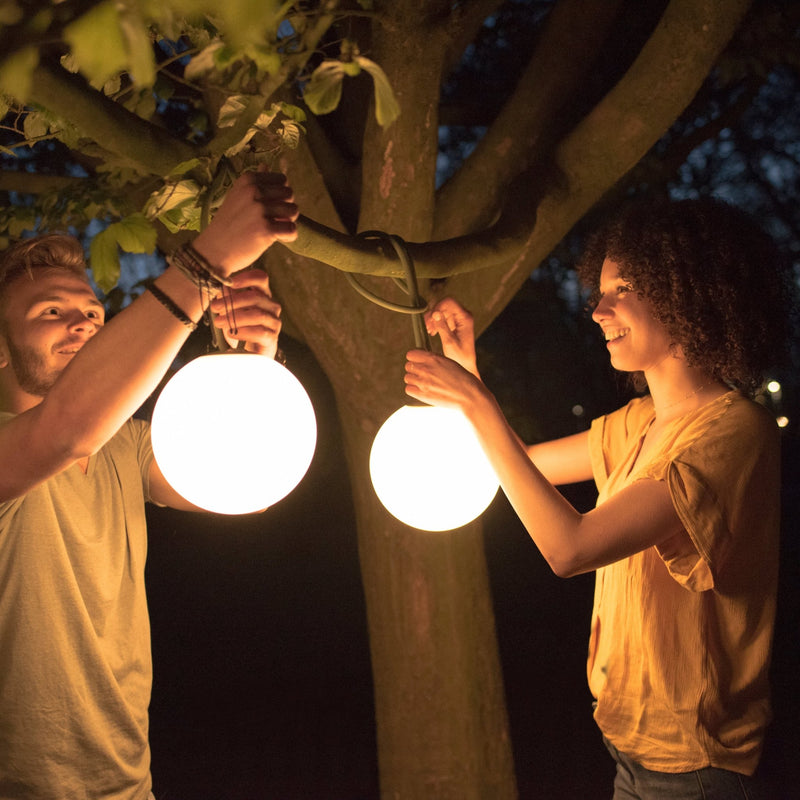 Deux personnes accrochent des lampes sans fil Fatboy Bolleke allumées dans un arbre, le soir